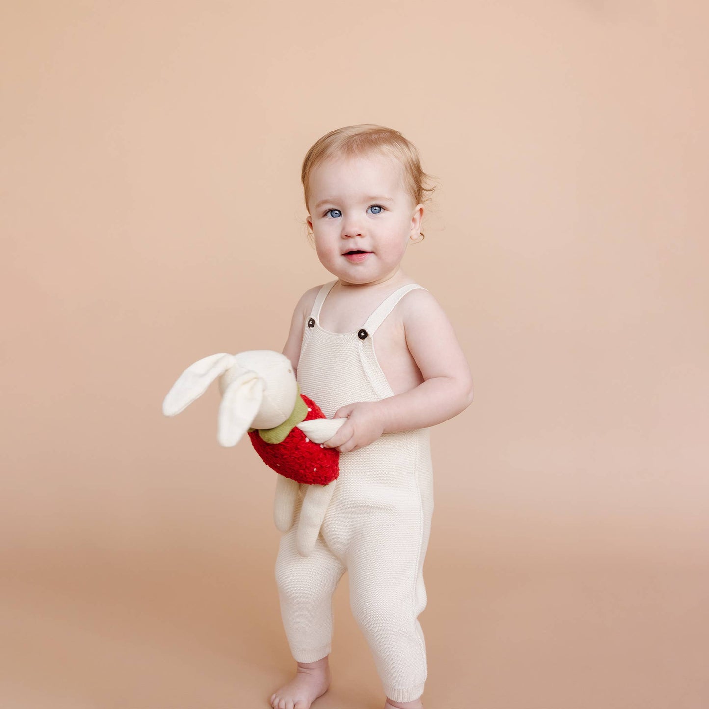 Baby in white overalls holding a bunny plush toy against a beige background