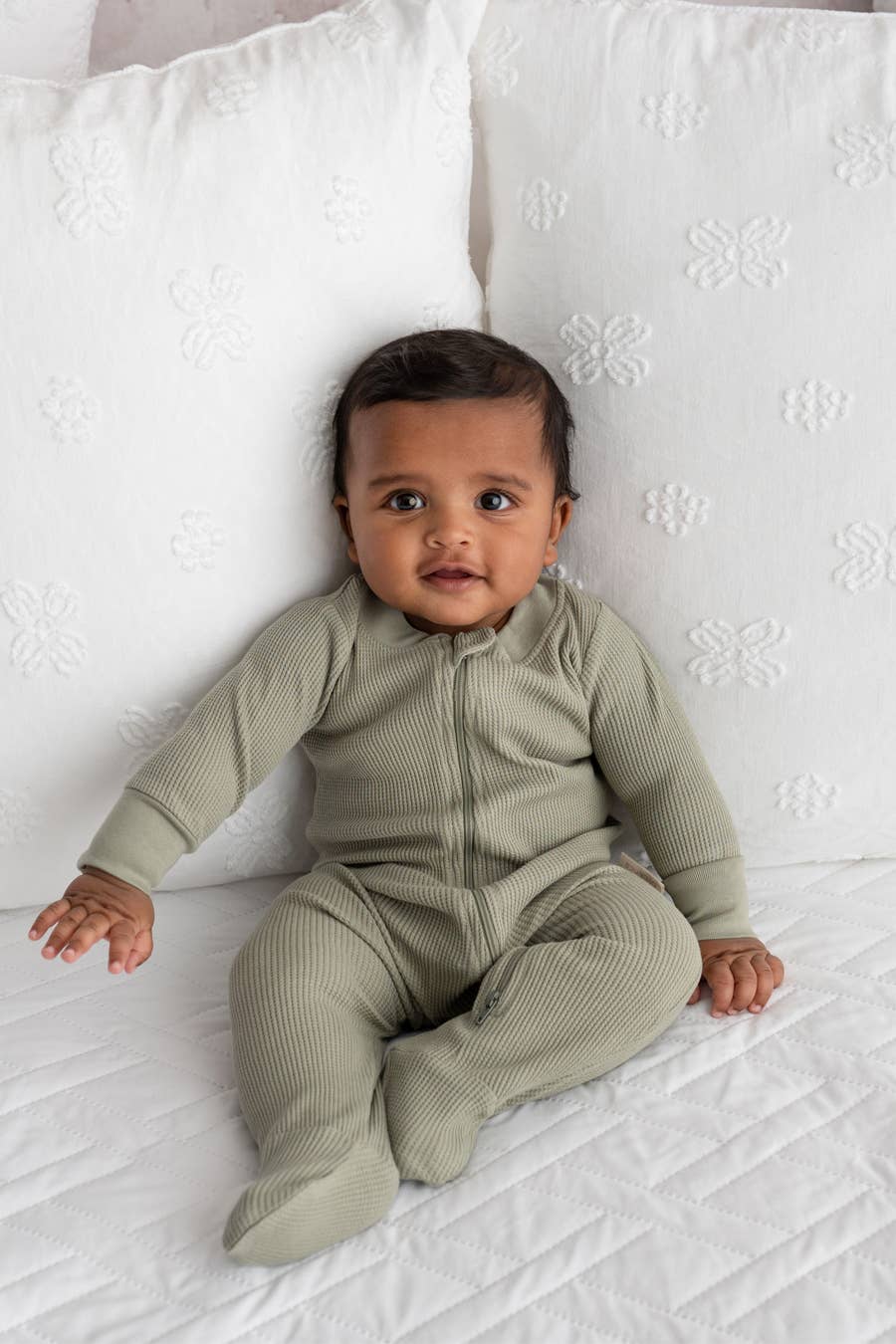 baby sitting on a bed wearing olive zip footie