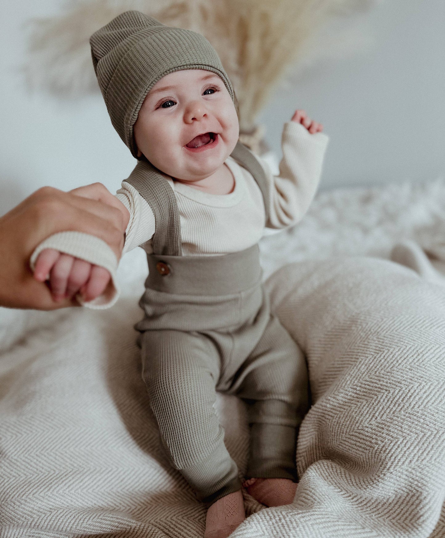 baby being held up on a bed wearing an organic waffle outfit with olive tights with suspenders