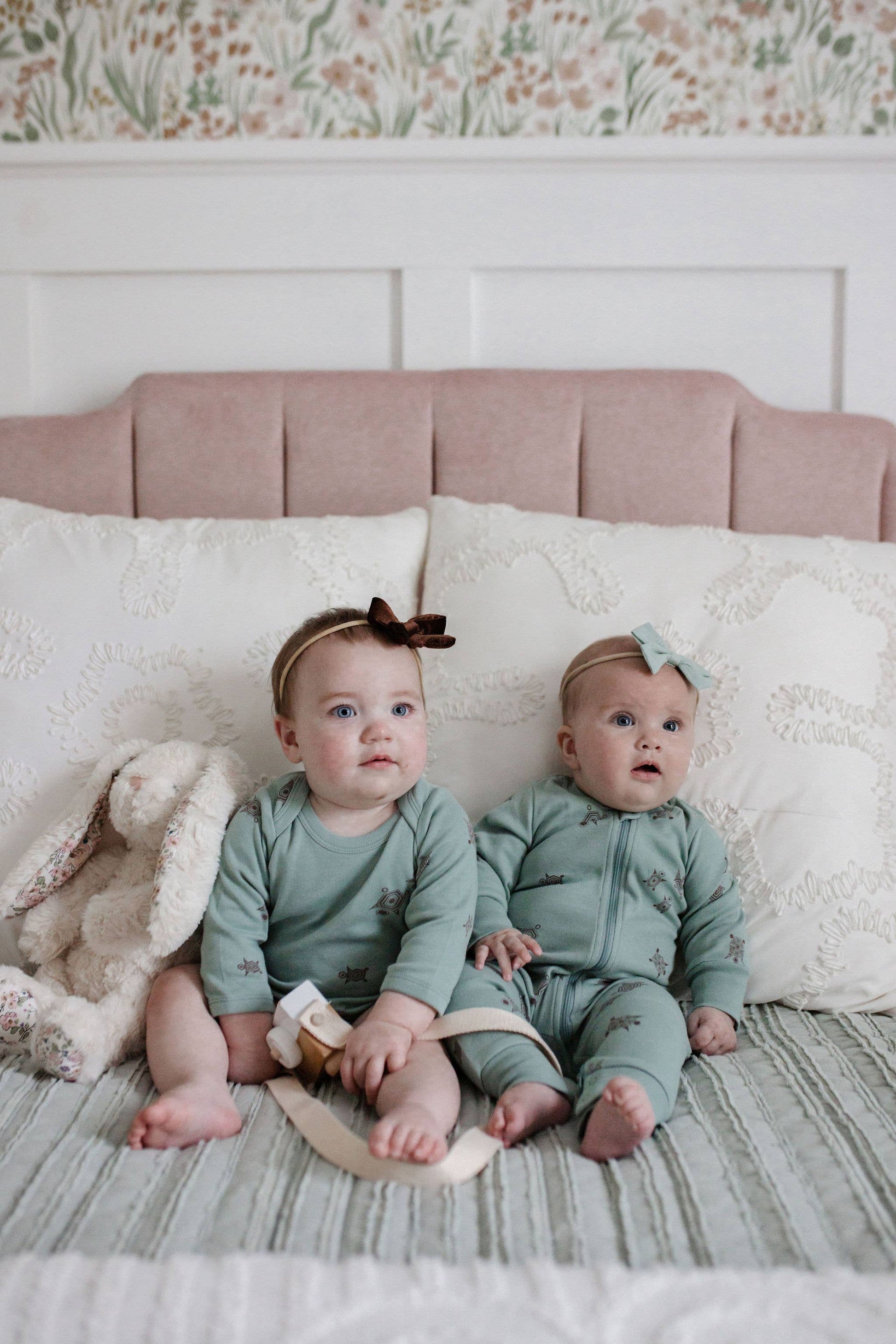 two babies sitting on a bed with a stuffed bunny wearing teal turtle print outfits