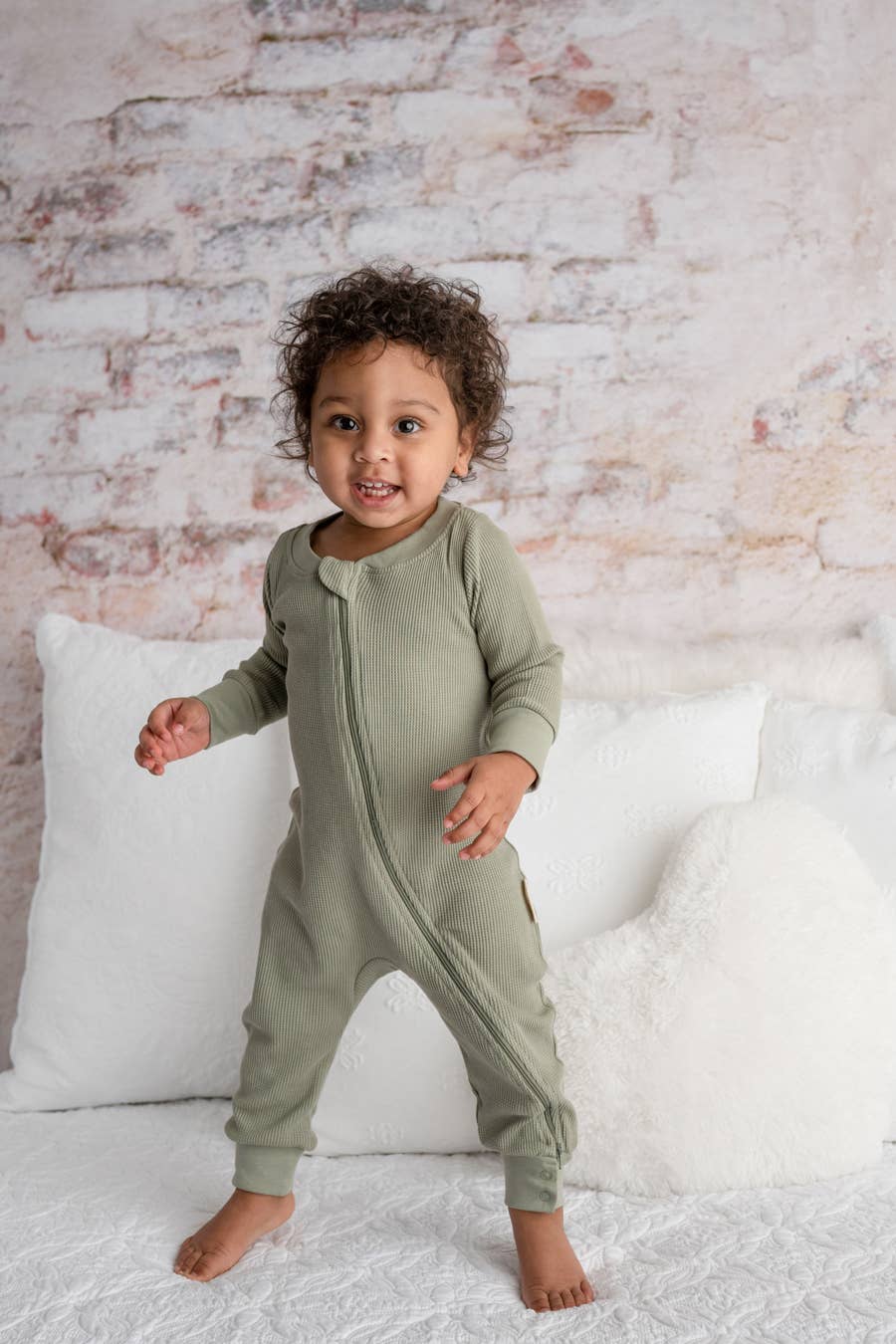 baby standing on a bed wearing organic waffle romper in olive