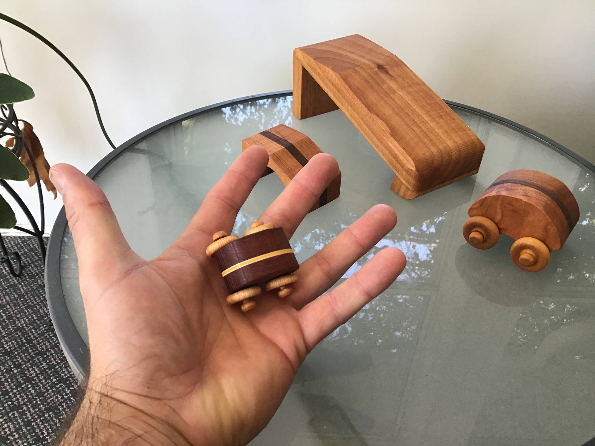 Hand holding a small wooden toy car with more wooden toys on a glass table.