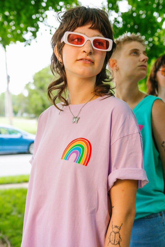 a person wearing pink rose-colored glasses and a pink t-shirt with a rainbow on the chest while standing outdoors