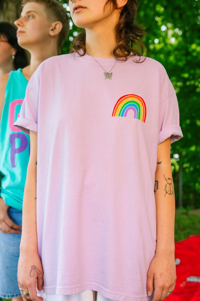 a person wearing a butterfly necklace and a pink t-shirt with a rainbow on the chest while standing outdoors