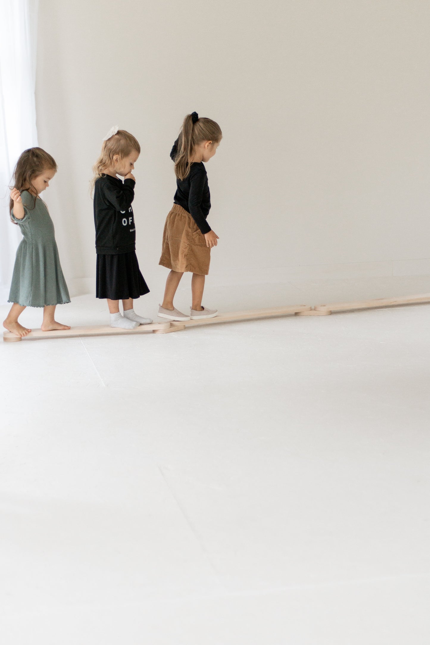 Three children standing on a wooden balance beam against a white wall.