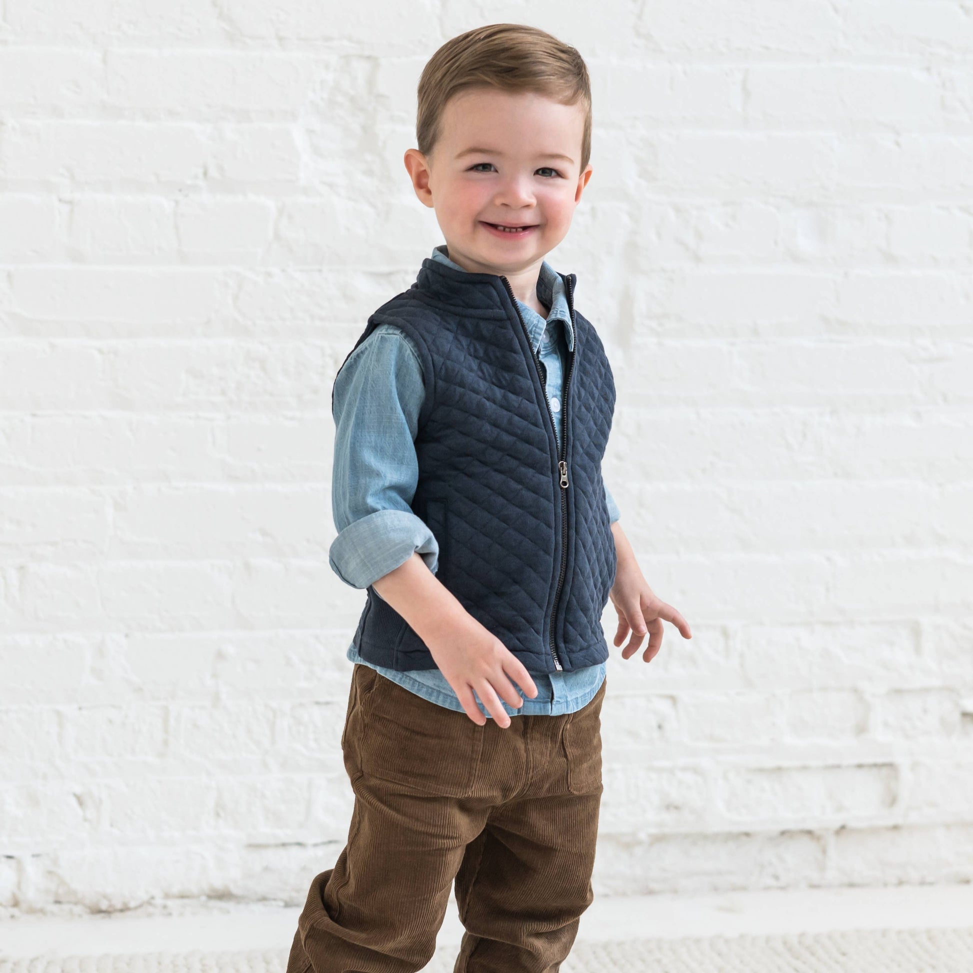 child standing in front of a white wall wearing a denim shirt, a blue zipper vest and sepia corduroy pants by colored organics