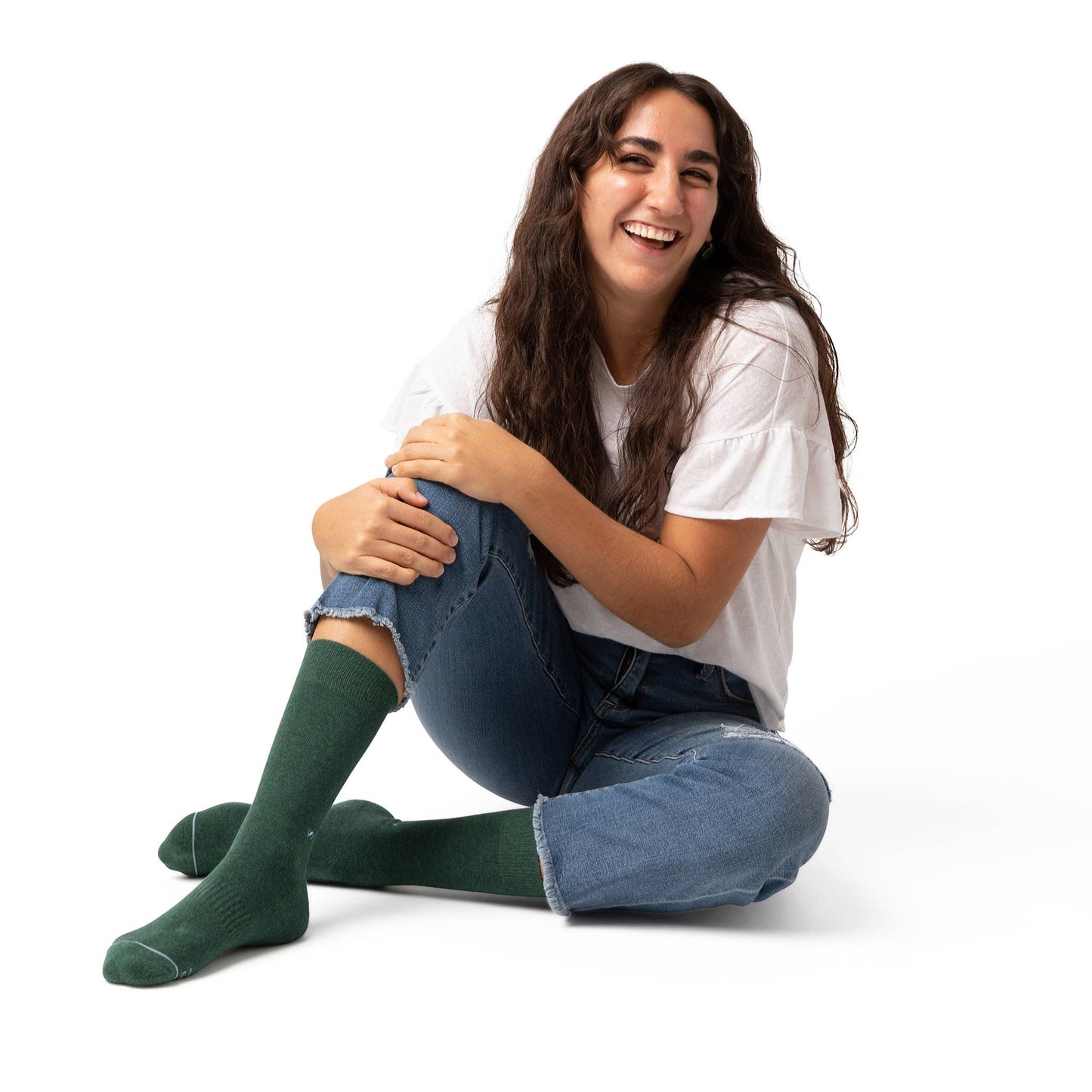 A person with long brown hair, jeans, and a white t-shirt sitting on the floor, laughing and wearing Socks that Plant Trees