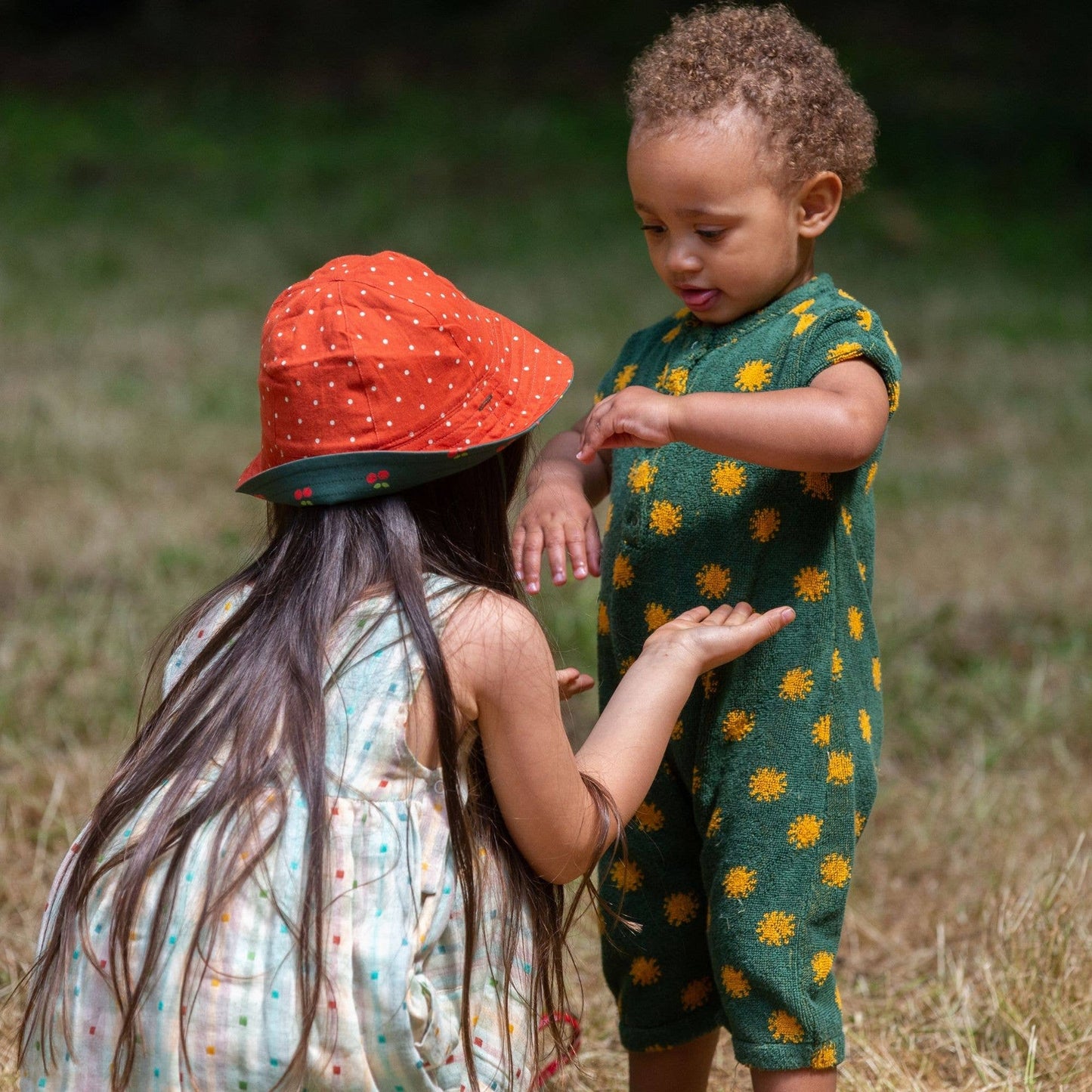 Two children playing outside in a grass field, one wearing Cherries Reversible Sunhat in Olive