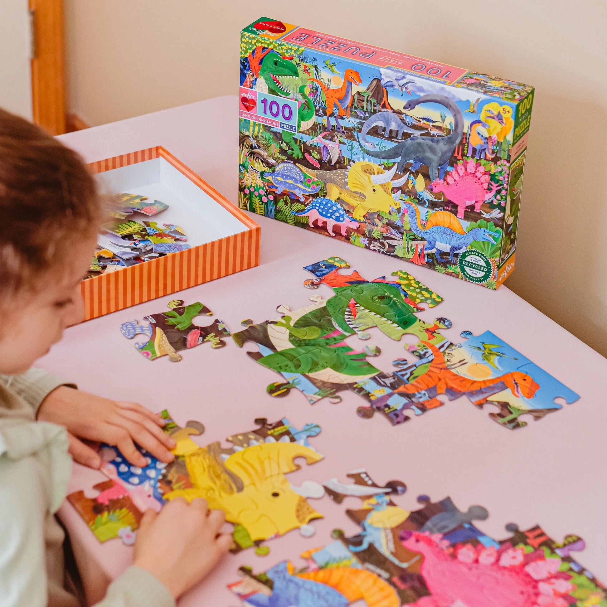 Child building a dinosaur-themed puzzle on a table next to the puzzle box.