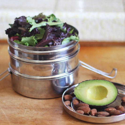 Metal 3-tier lunch container with salad, almonds and half an avocado on a wooden surface
