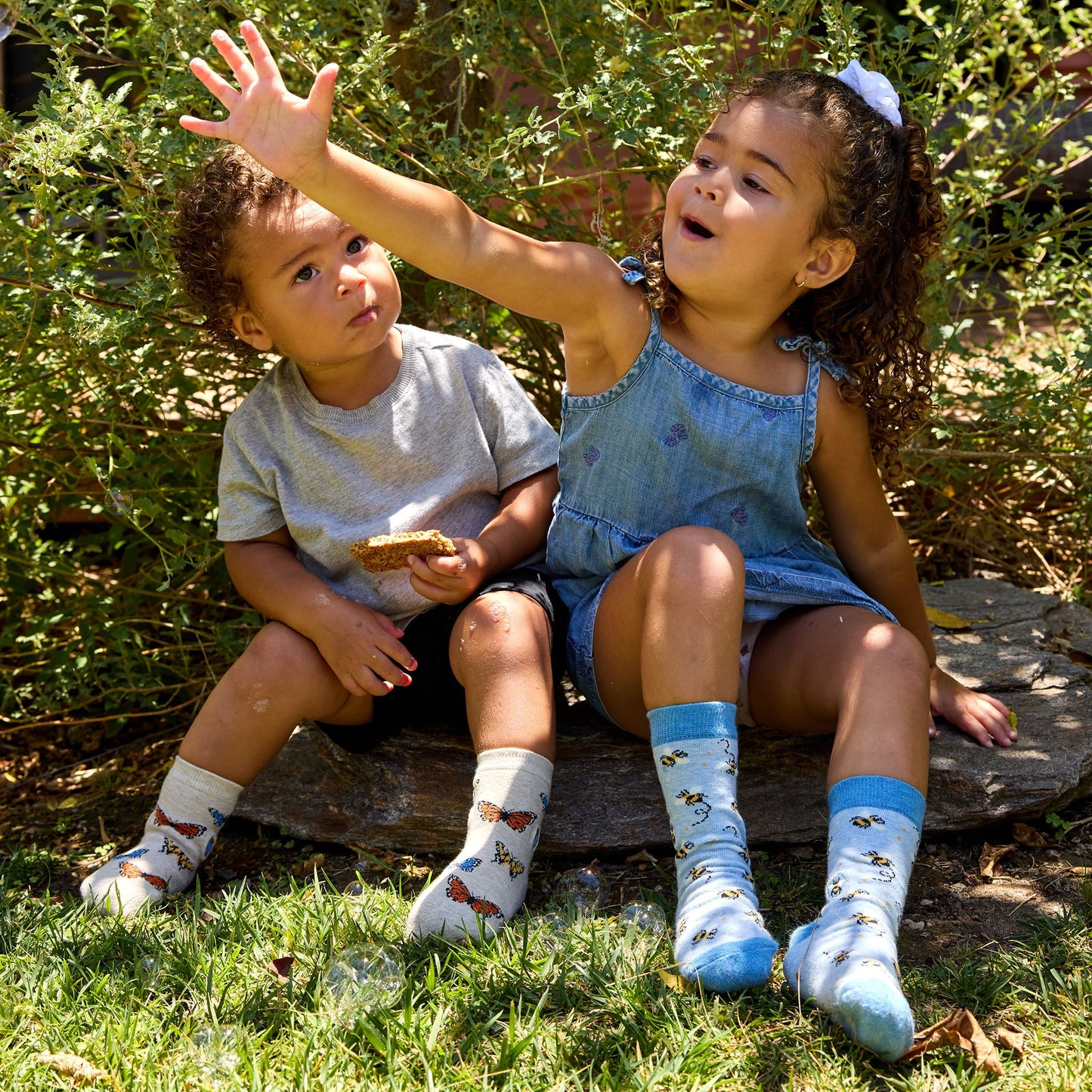 Two children sitting on a rock outdoors, surrounded by greenery.