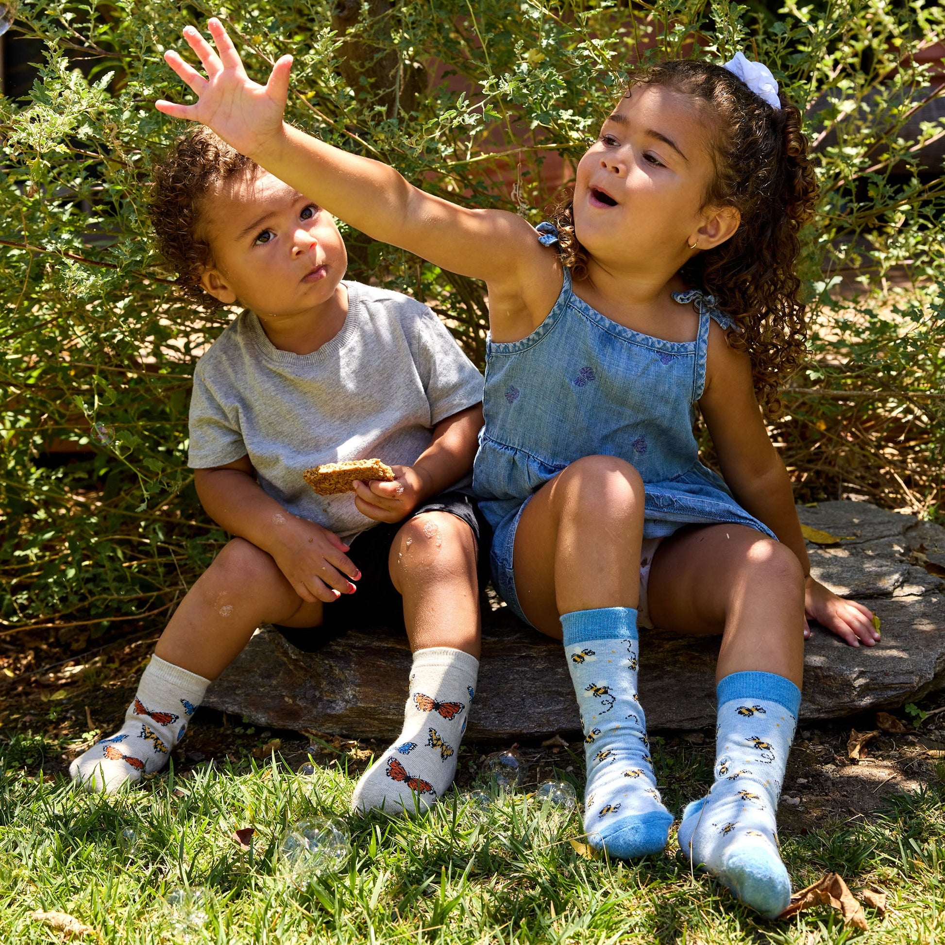 Two children sitting on a rock outdoors, surrounded by greenery.