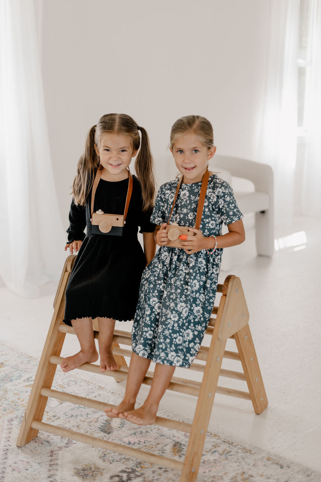 Two young girls standing on a wooden step stool in a bright room.