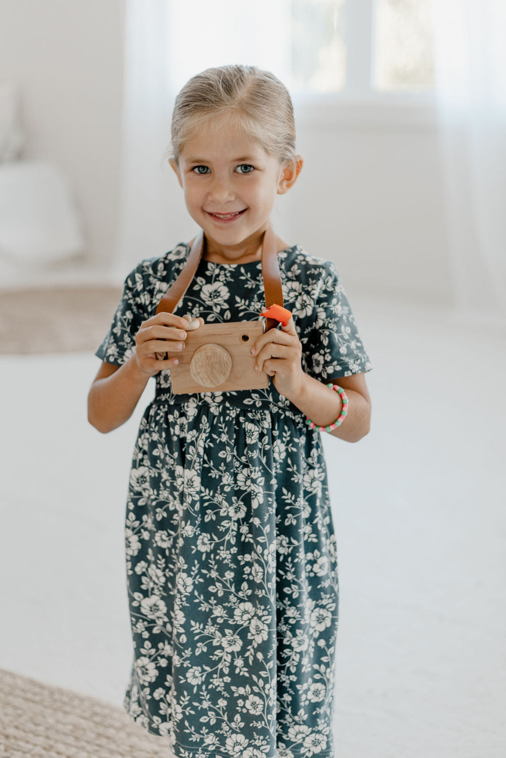 Young girl in a floral dress holding a toy camera indoors.
