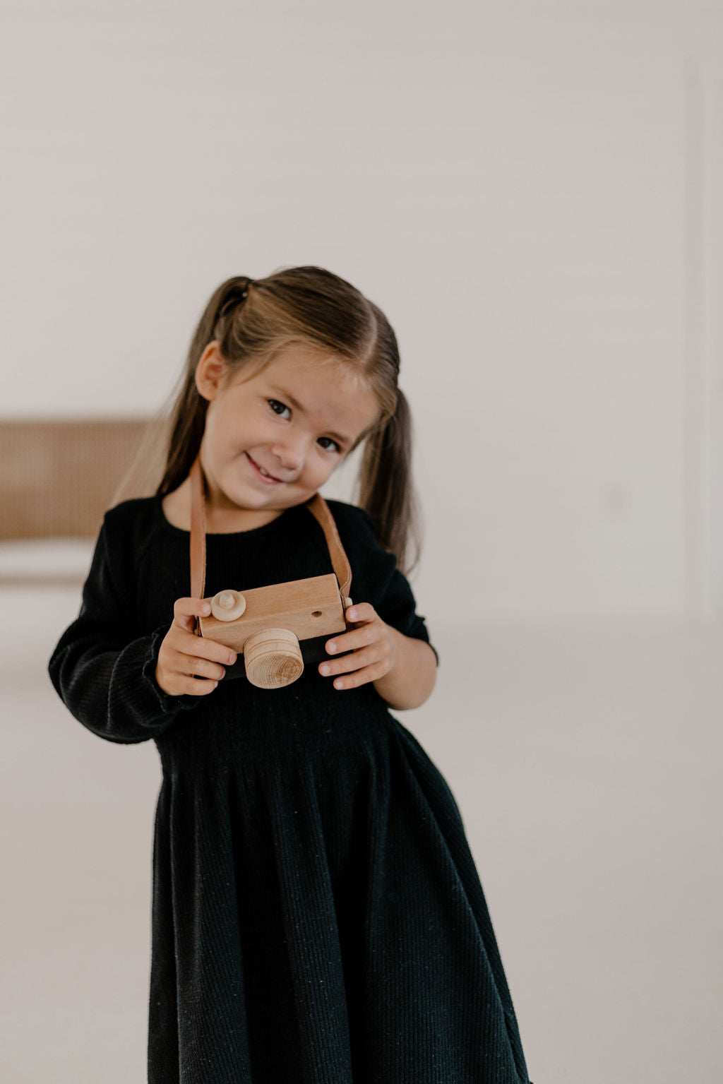 Young girl in a black dress holding a wooden toy camera against a plain background