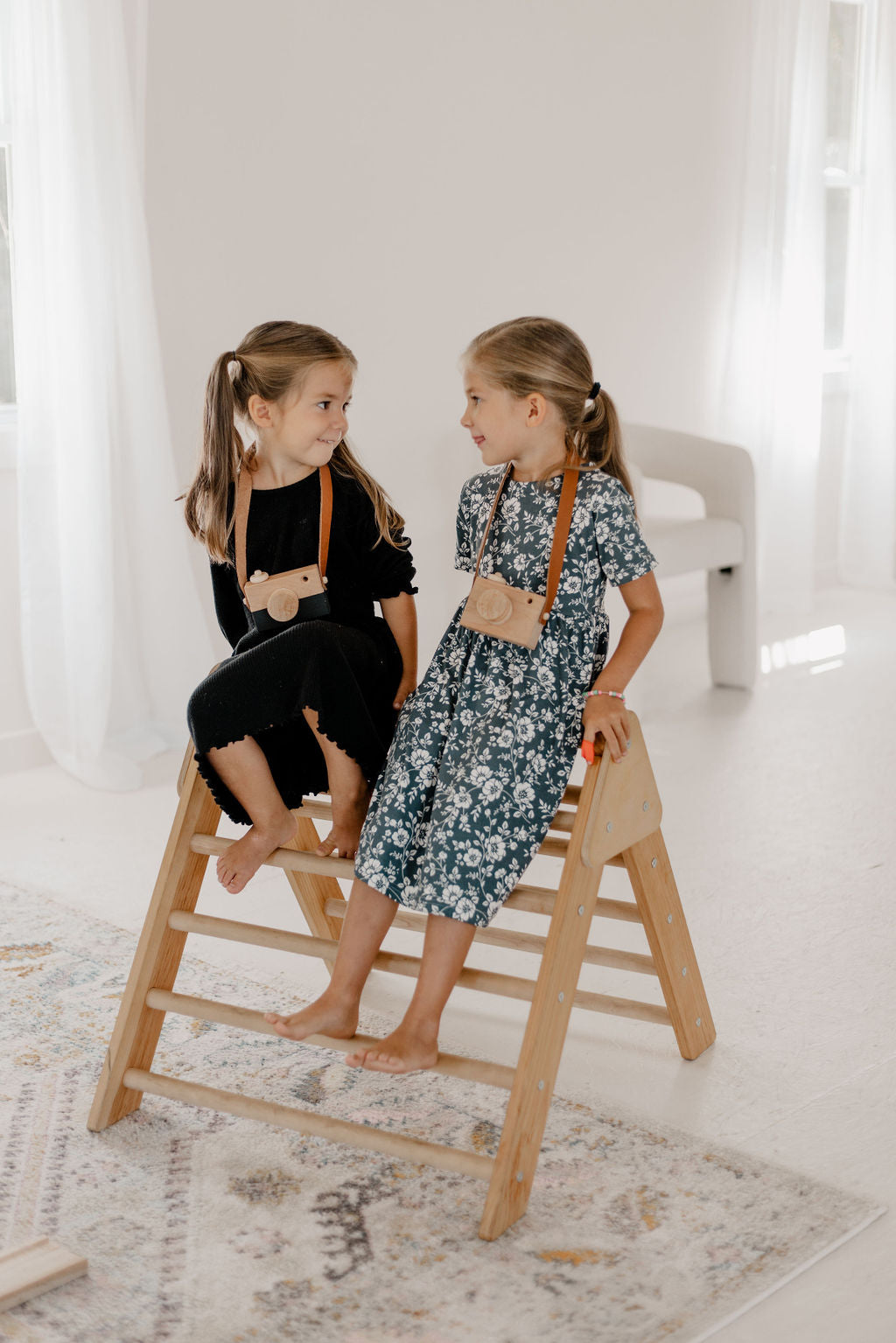 Two young girls sitting on a wooden stool in a bright room.