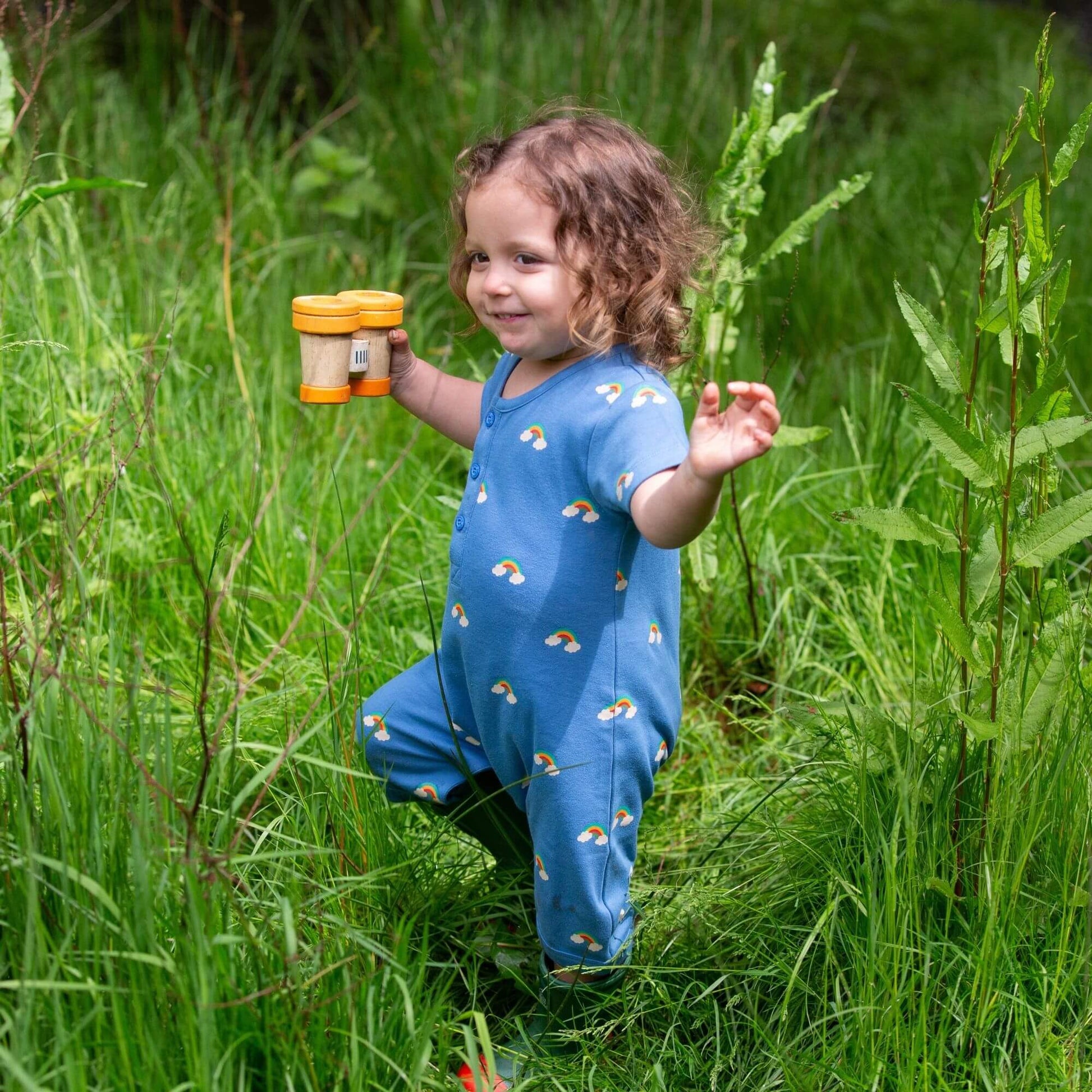 a baby walking around in tall grass wearing a blue rainbows summer romper and green rain boots, holding toy binoculars