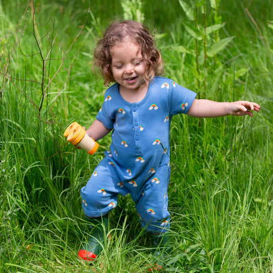 a toddler running in the grass and laughing, wearing a blue rainbow romper and green rain boots