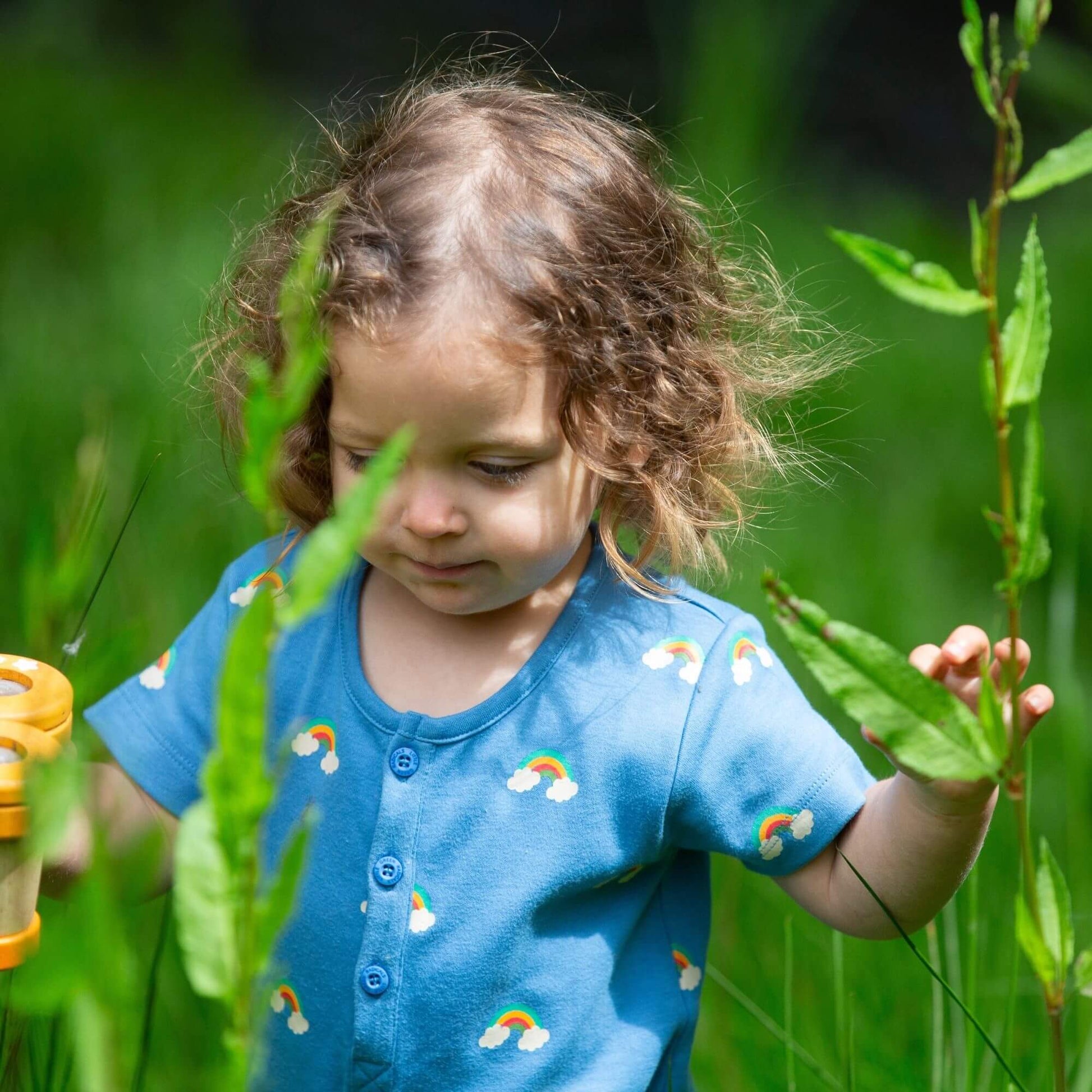 a baby standing in tall grass wearing a blue rainbows summer romper holding toy binoculars