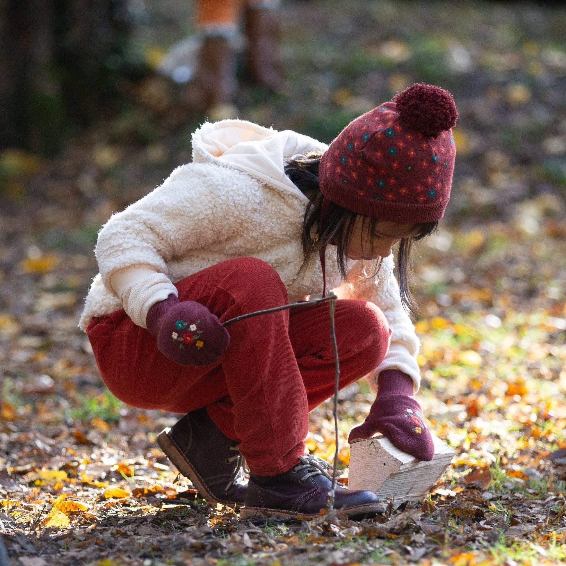 Child crouching on forest ground holding a stick wearing sherpa fleece campfire jacket