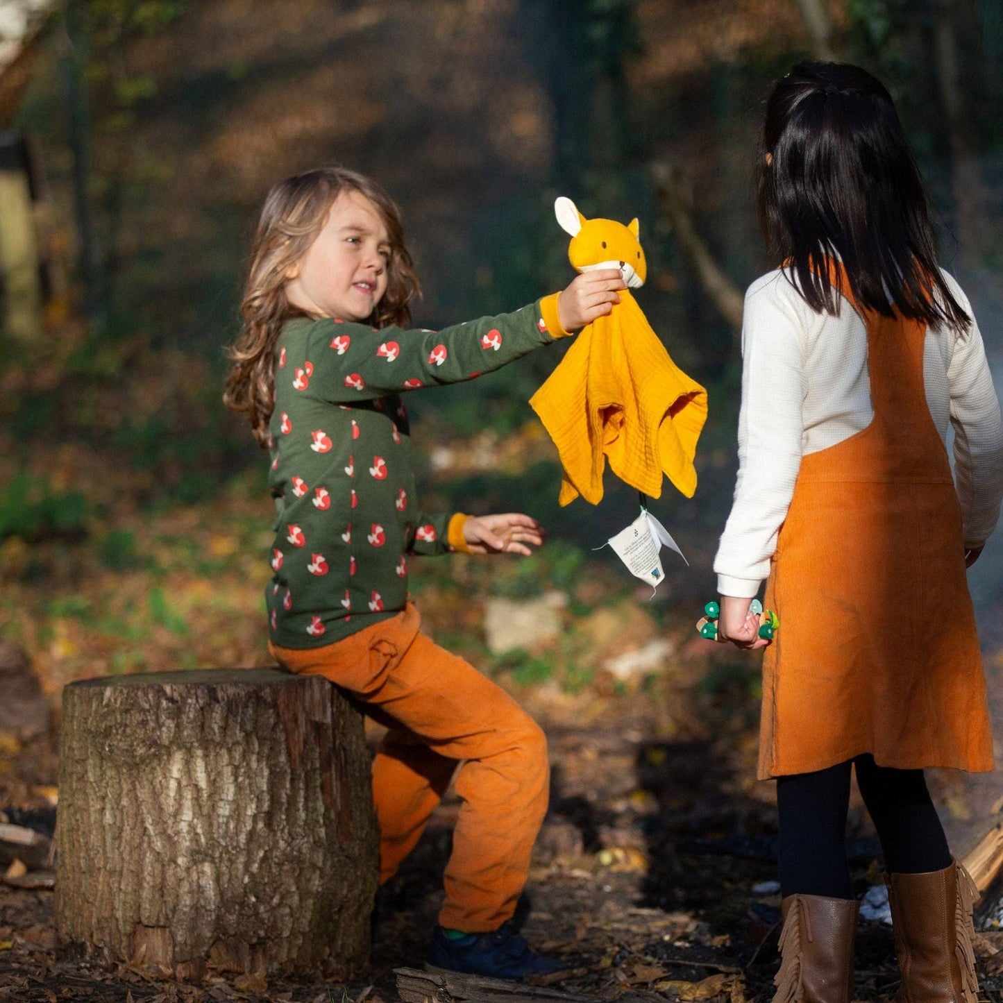 a child sitting on a log in the woods holding an organic cotton fox lovey