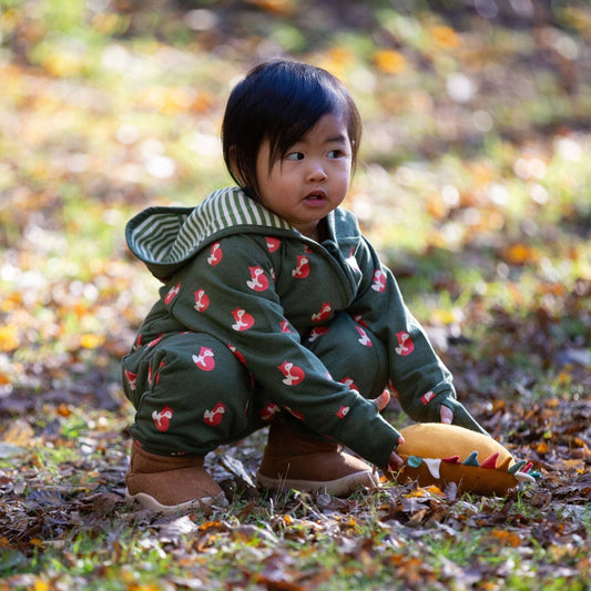 a toddler crouching on the ground outside wearing a foxes snug-as-a-bug suit and holding a sun soft stuffed toy