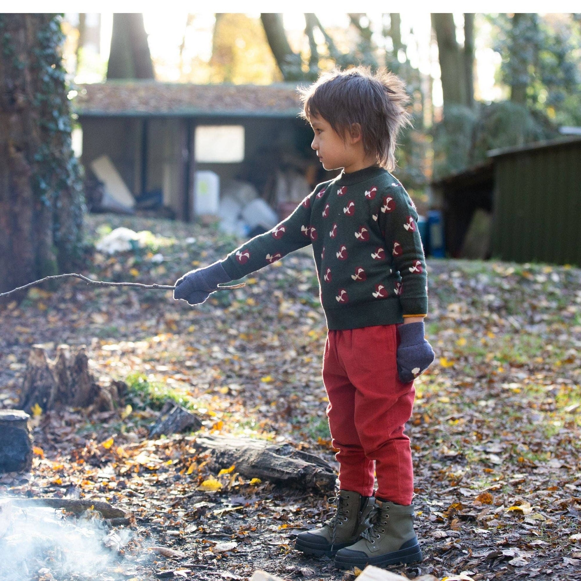 child standing in the woods holding a stick wearing little foxes knitted sweater