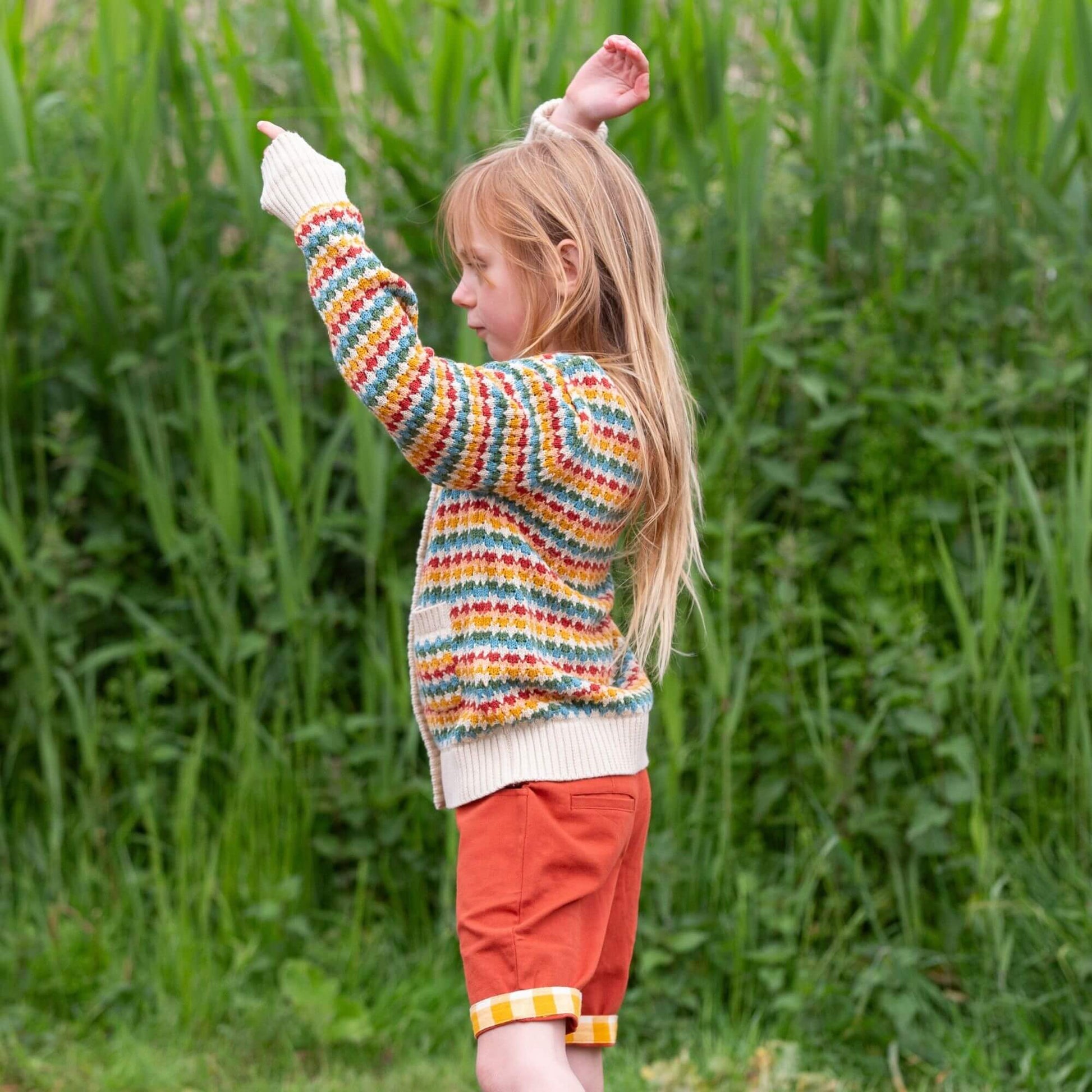 a child standing outside wearing red shorts and organic rainbow knitted cardigan