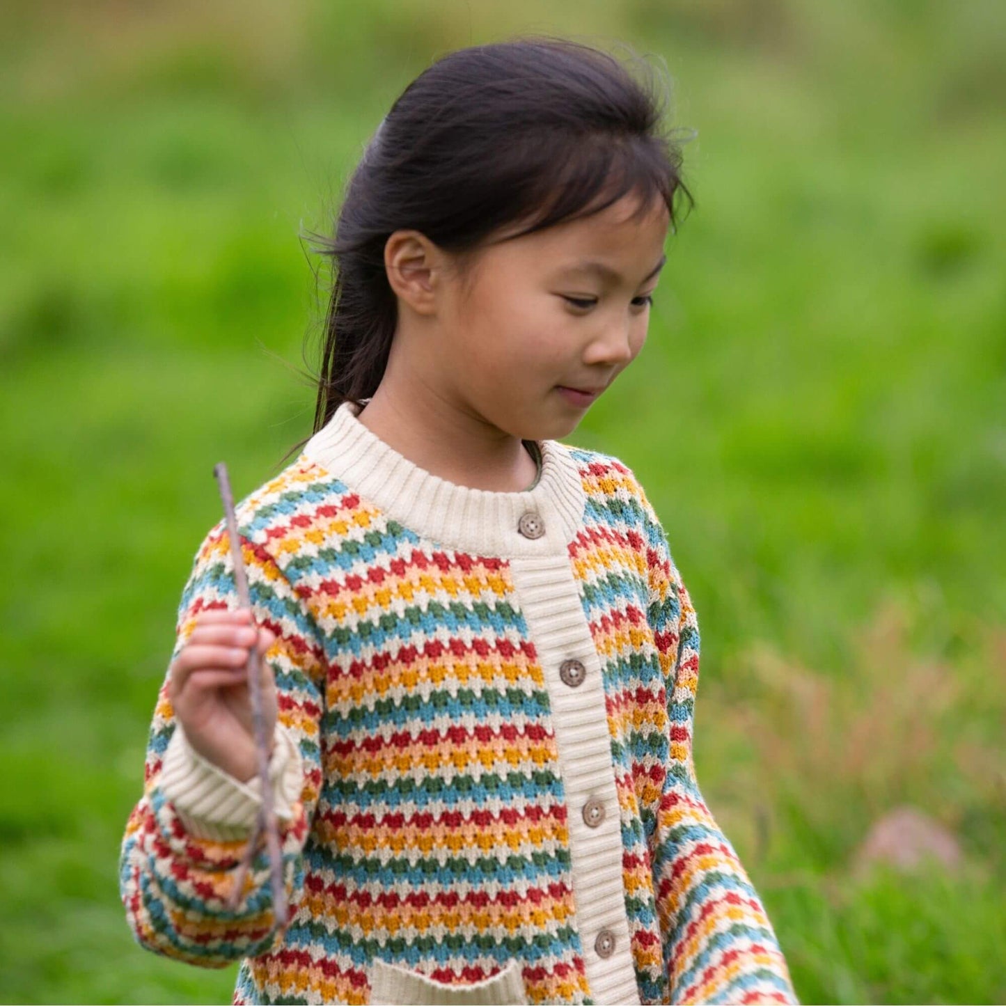 a child standing outdoors holding a stick wearing a knitted organic rainbow striped cardigan