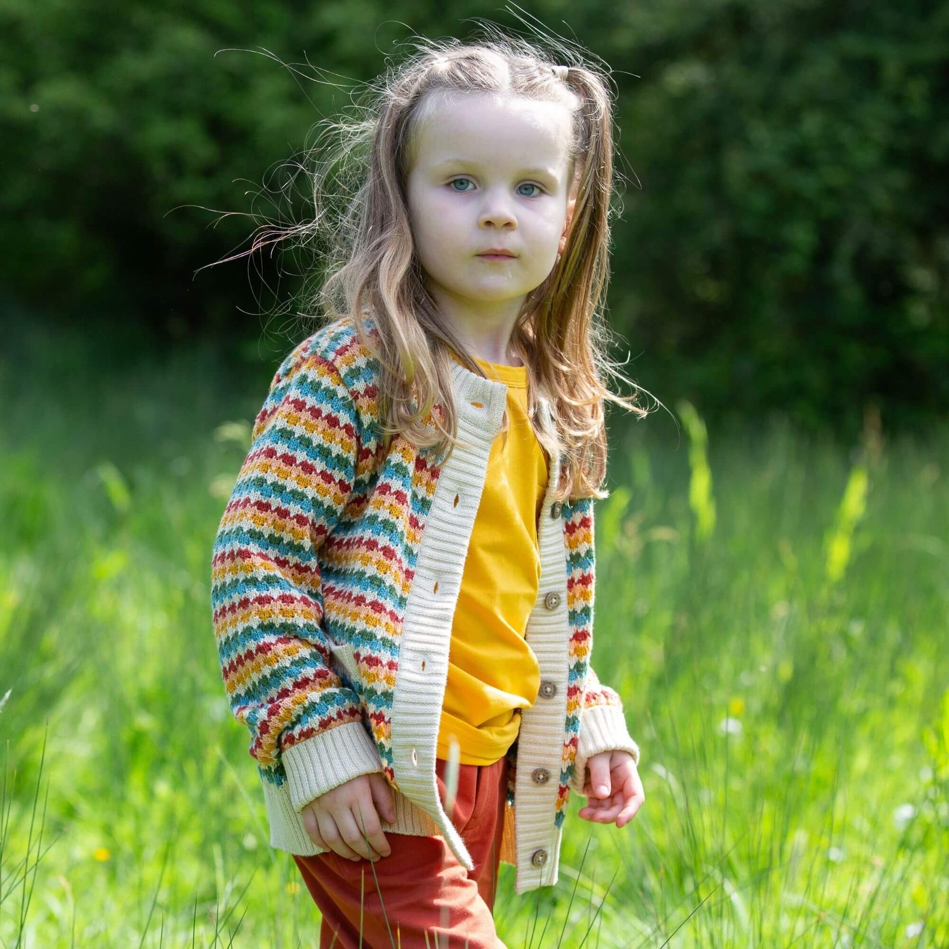 a child standing in the grass wearing red shorts, a yellow t-shirt, and a rainbow striped organic knitted cardigan