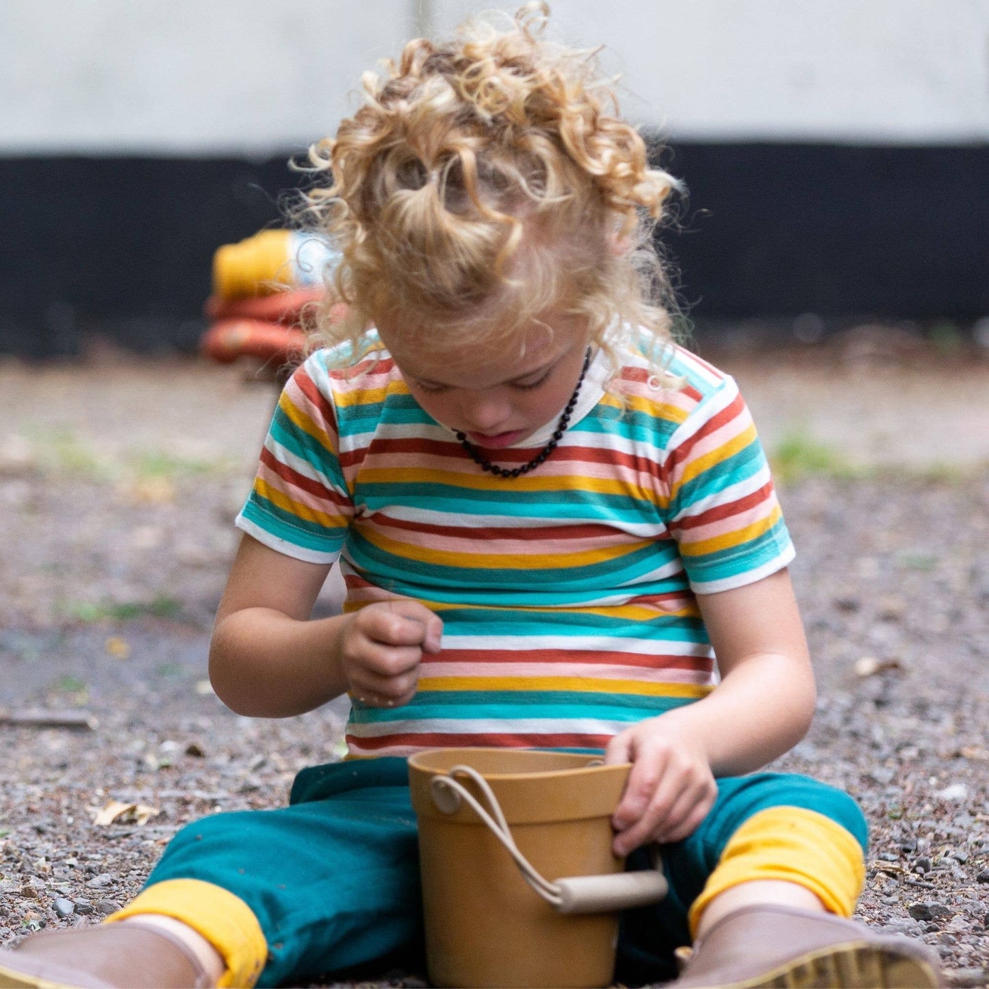 a child sitting outside filing a bucket with dirt wearing a rainbow striped short sleeve tee and blue joggers