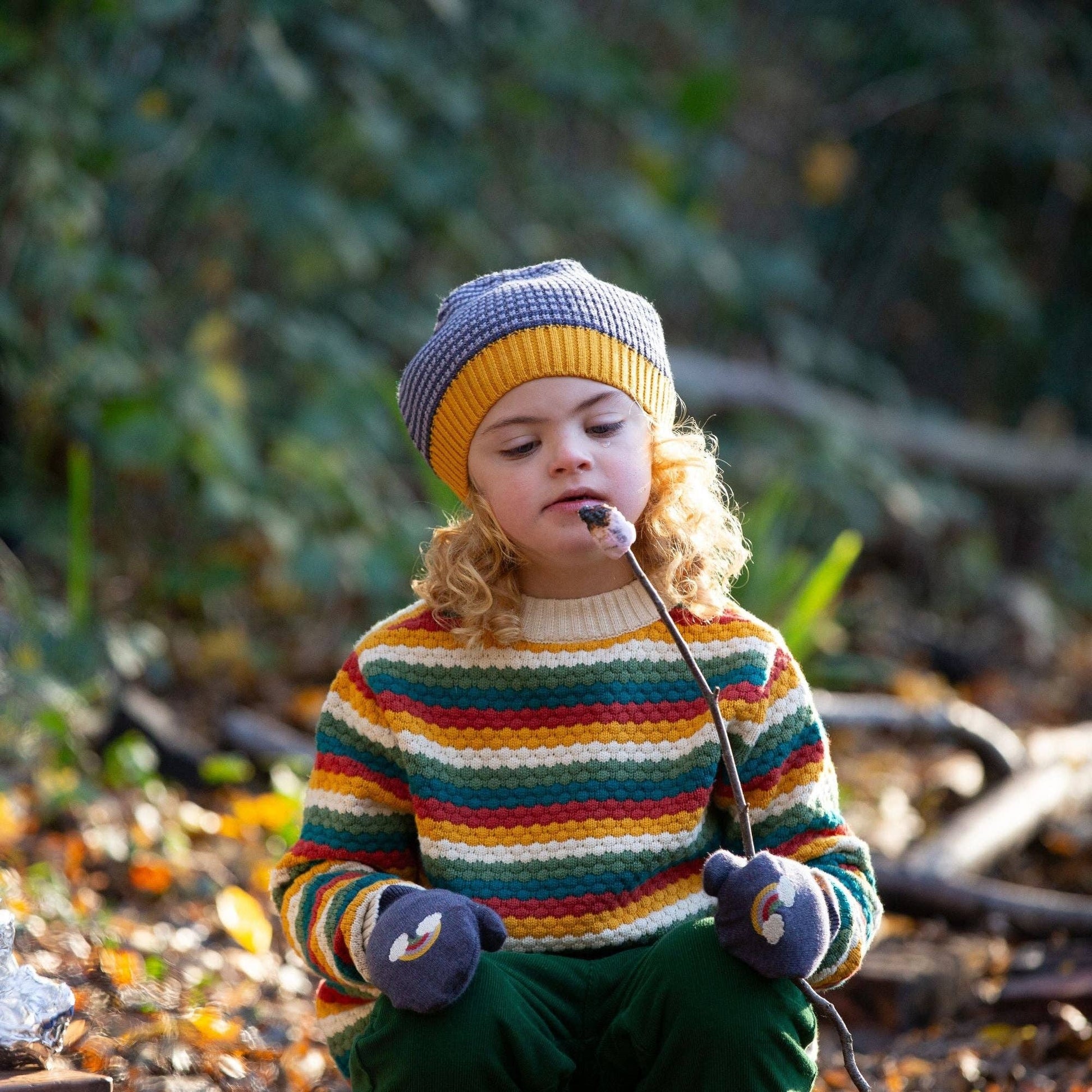 A child sitting outdoors eating a s'more wearing a rainbow stripe knitted sweater