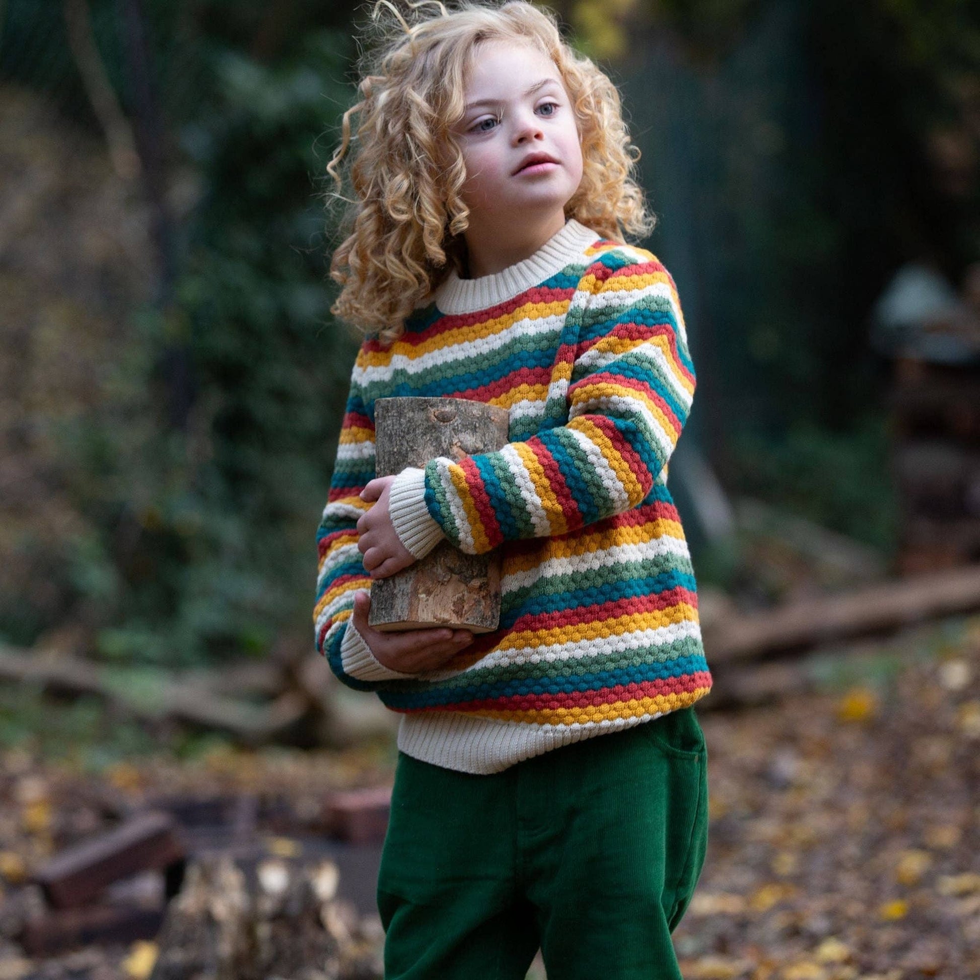 Child standing in the woods holding a small tree log wearing organic rainbow stripes knitted sweater