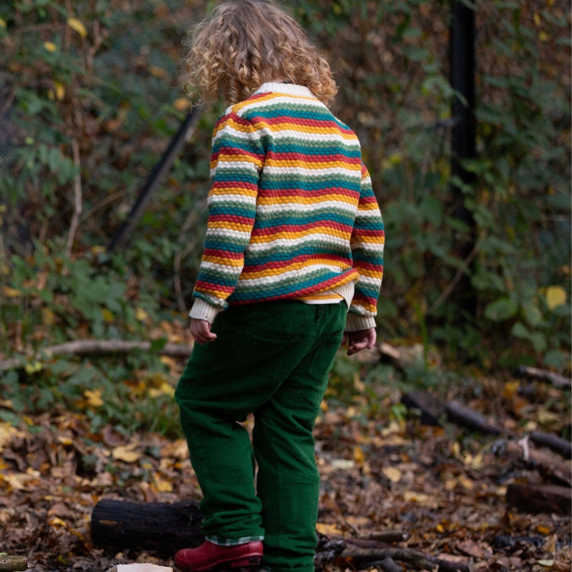 a child standing in the woods with their back to the camera wearing a rainbow striped organic knitted sweater