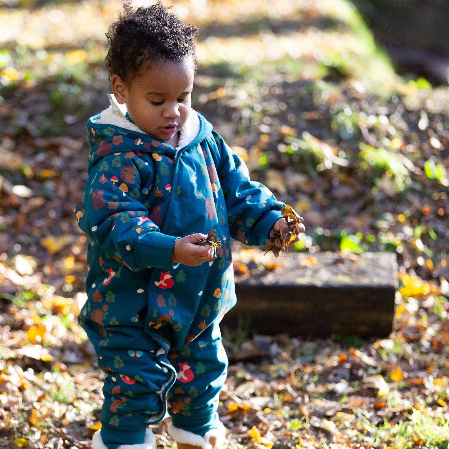 toddler standing outside holding handfuls of leaves wearing enchanted forest sherpa-lined snowsuit