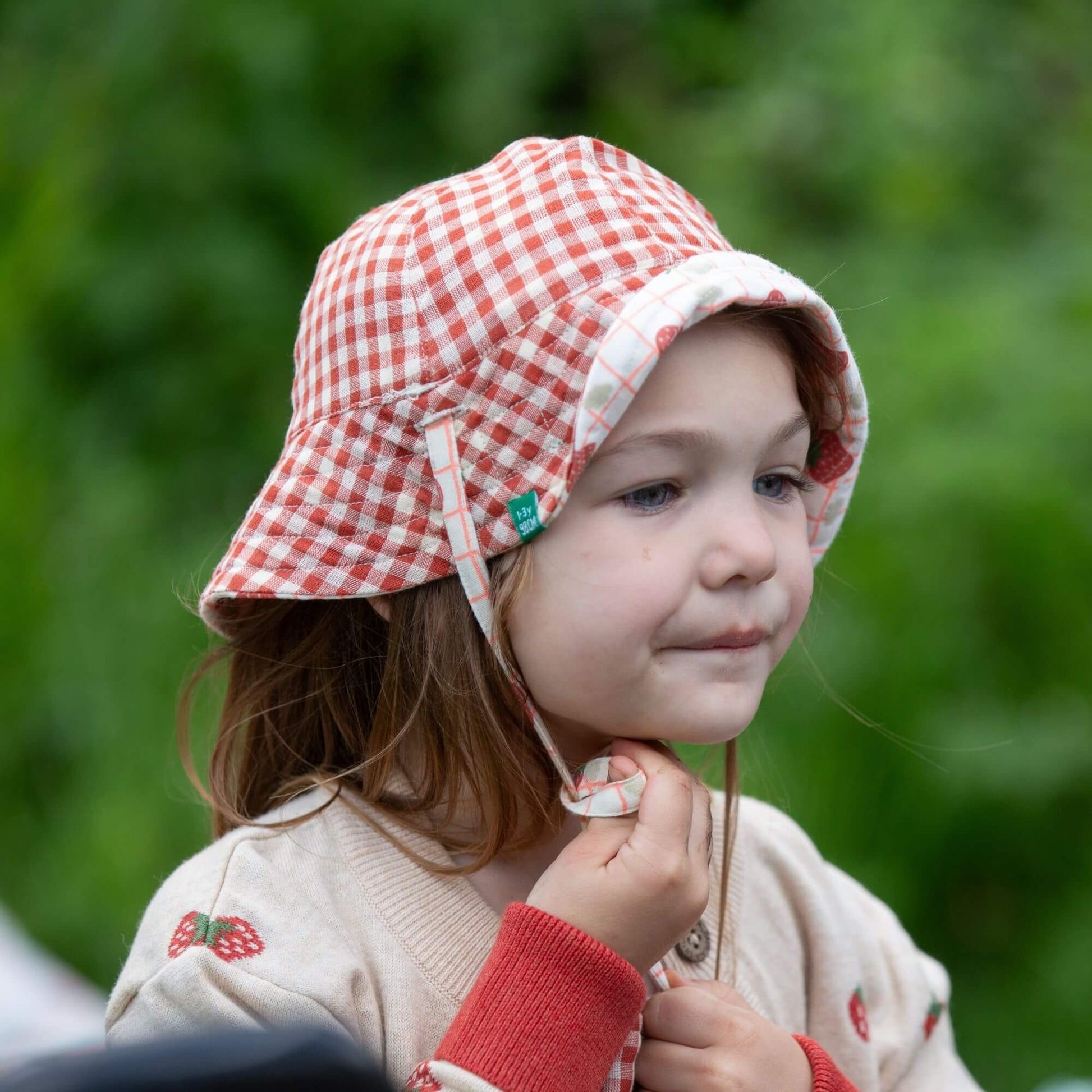 a child standing outdoors wearing a strawberries knitted cardigan and tying an organic reversible strawberries hat nder their chin