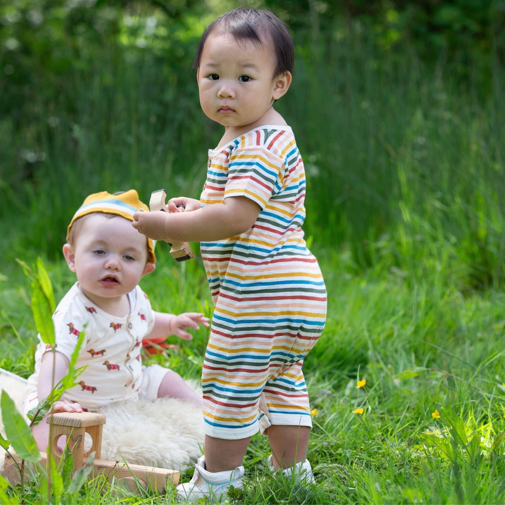 a baby standing outdoor wearing a cream rainbow organic summer romper and holding a wooden toy, and another baby sitting in the grass wearing a summer romper playing with wooden blocks