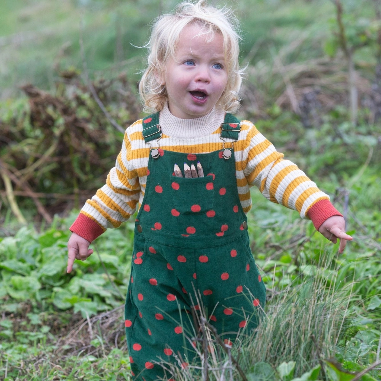 child running outside wearing a yellow and white striped sweater under apple printed corduroy overalls