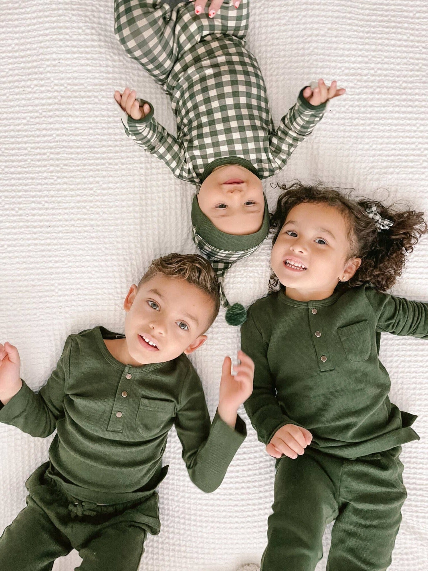 Two children wearing green thermal lounge sets and a baby wearing a green checkered romper and matching hat lying on a textured white blanket
