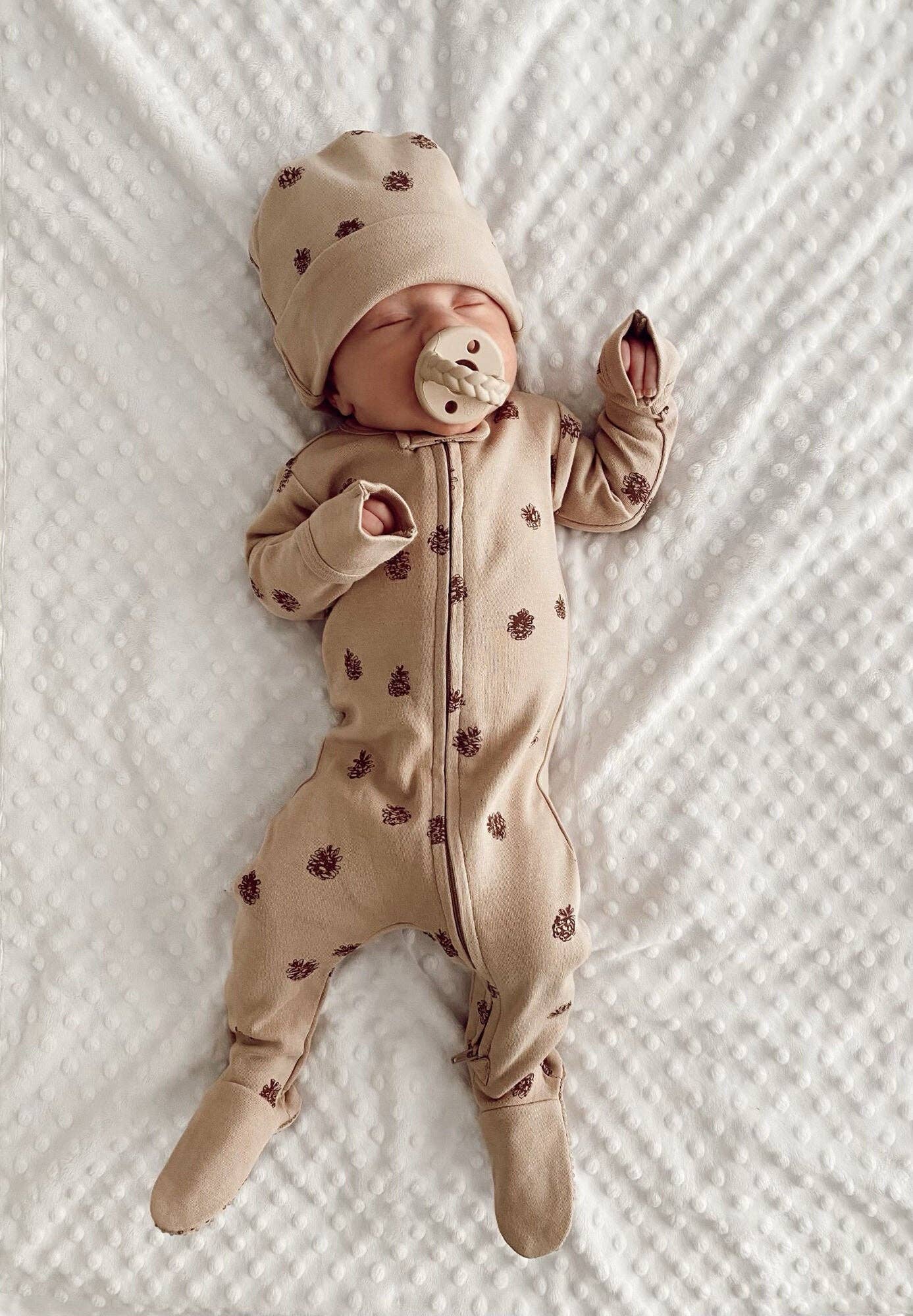 Baby in a beige pinecones romper and matching hat with a pacifier, lying on a textured white blanket.
