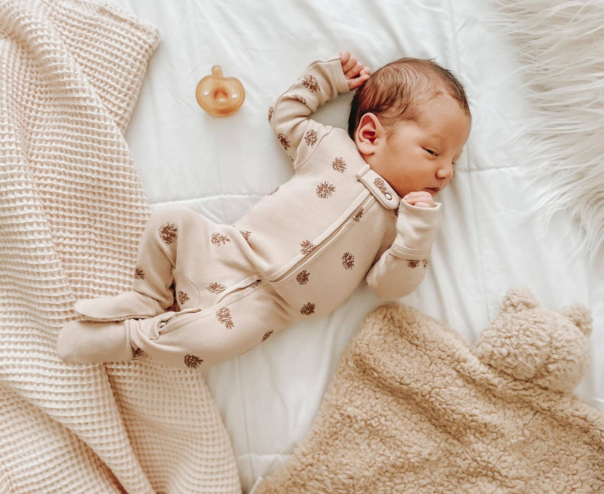 Newborn baby in a beige pinecones footie lying on a soft surface with a teddy bear lovey and a pacifier.