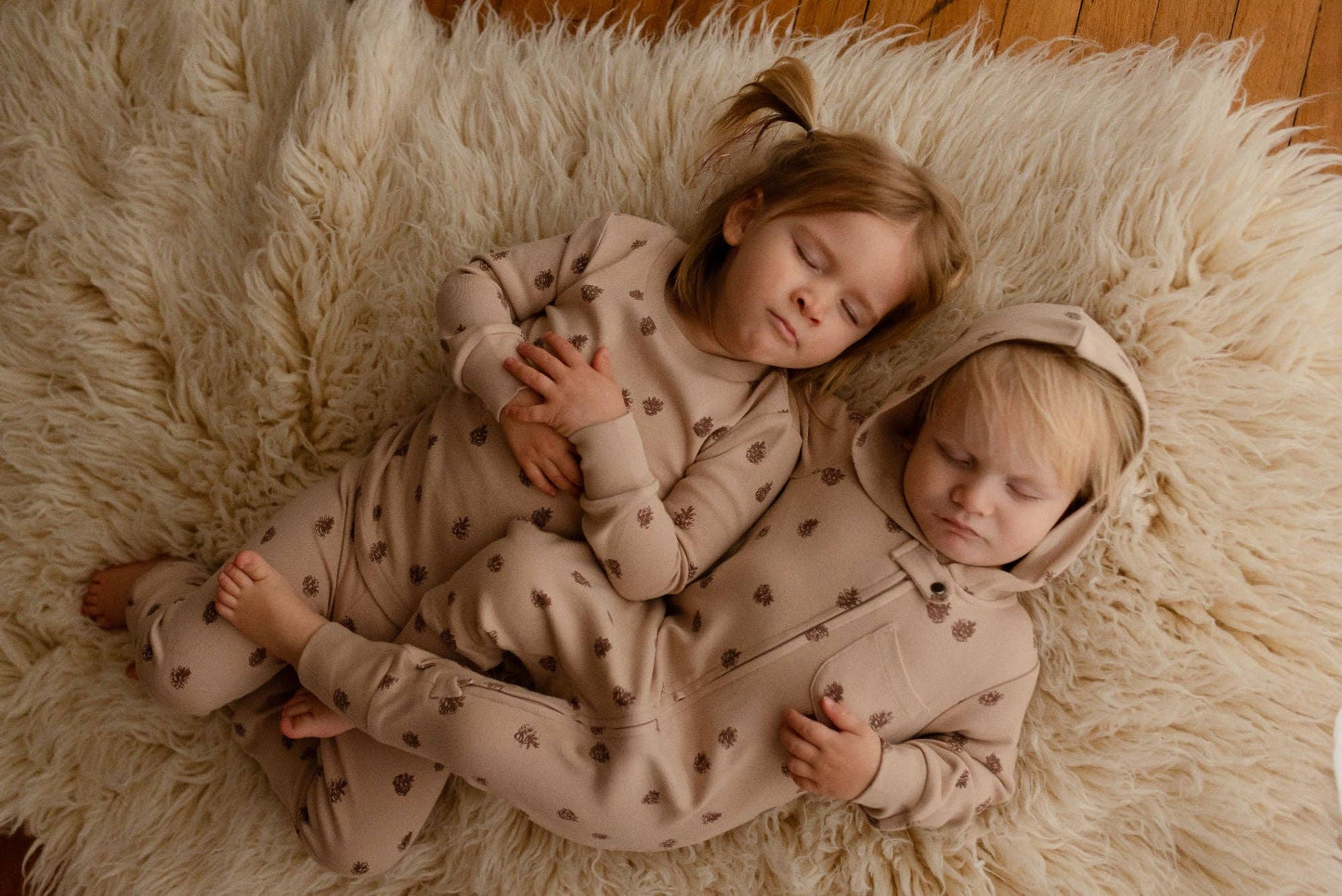Two children in matching pinecones pajamas lying on a fluffy white rug.
