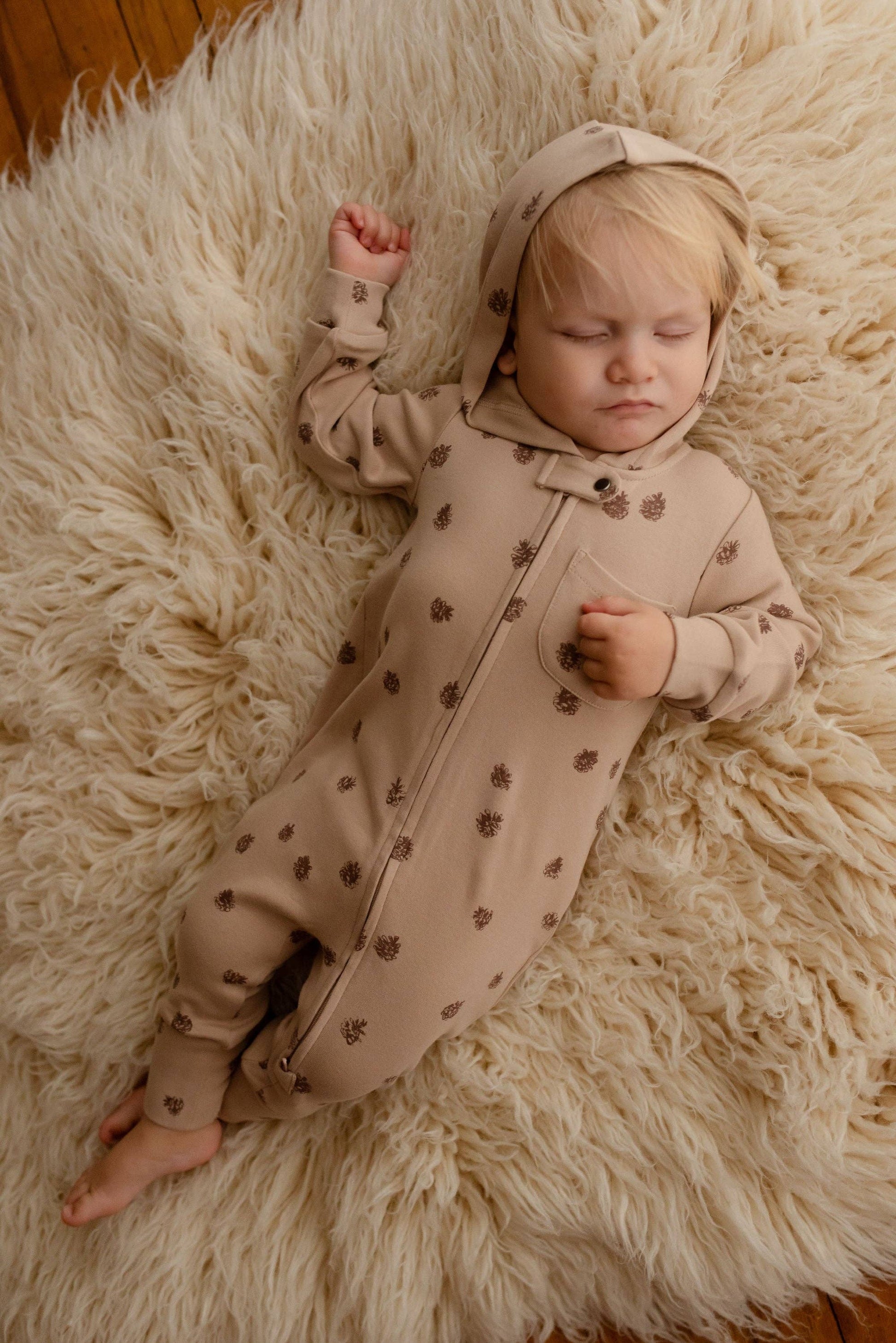 Baby in a beige romper with brown pinecones print lying on a fluffy white rug.