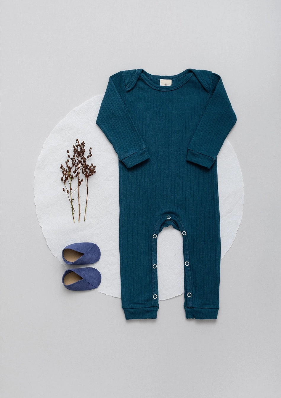 Long sleeve ribbed romper in petroleum blue laid flat next to some flowers and a pair of baby shoes