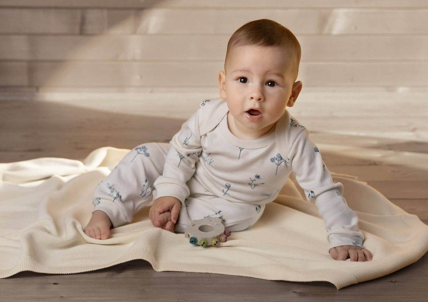 Baby sitting on a blanket on the floor with a wooden rattle wearing a plants delight romper