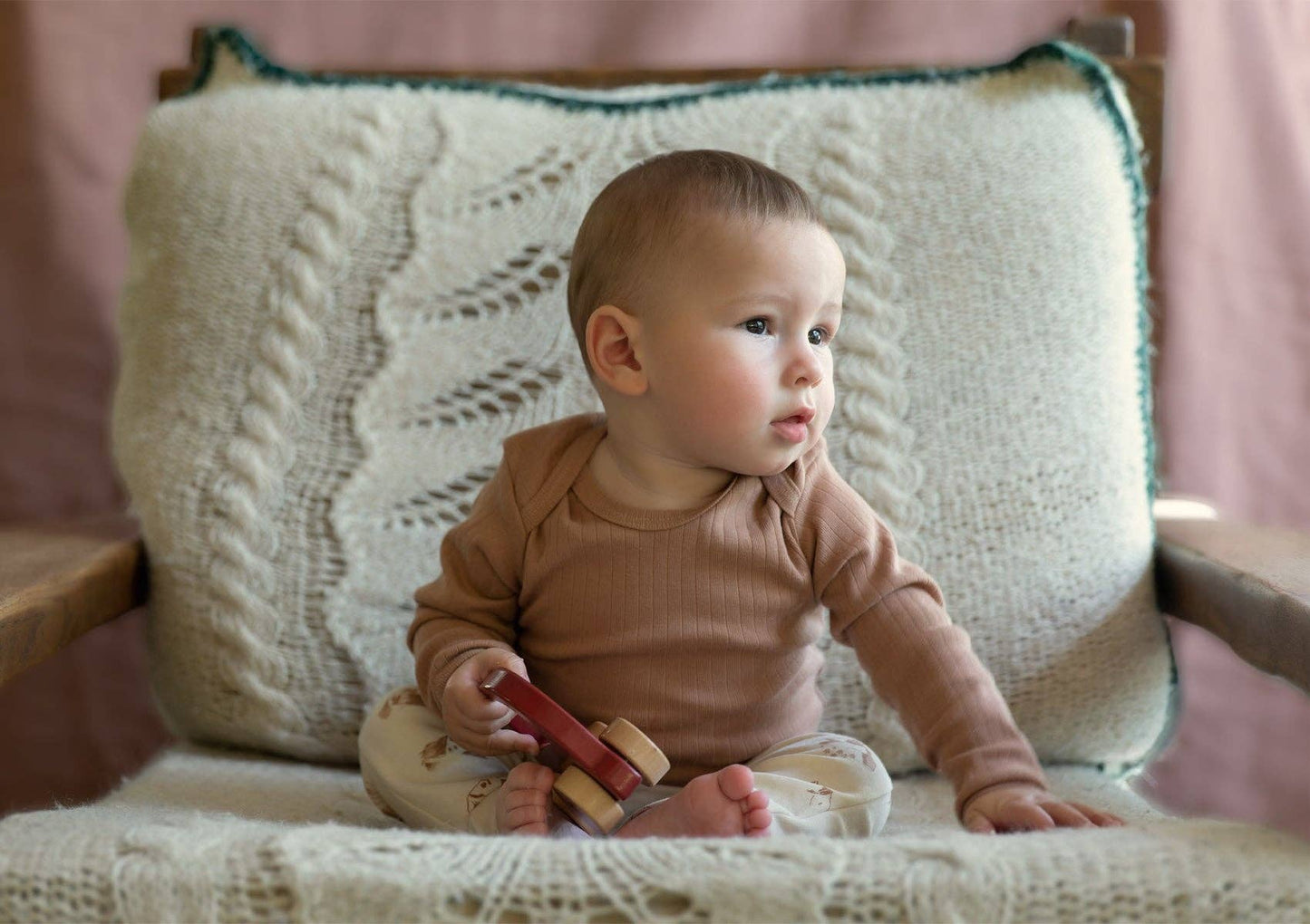 Baby sitting on a chair holding a wooden car wearing a sienna long-sleeve ribbed onesie