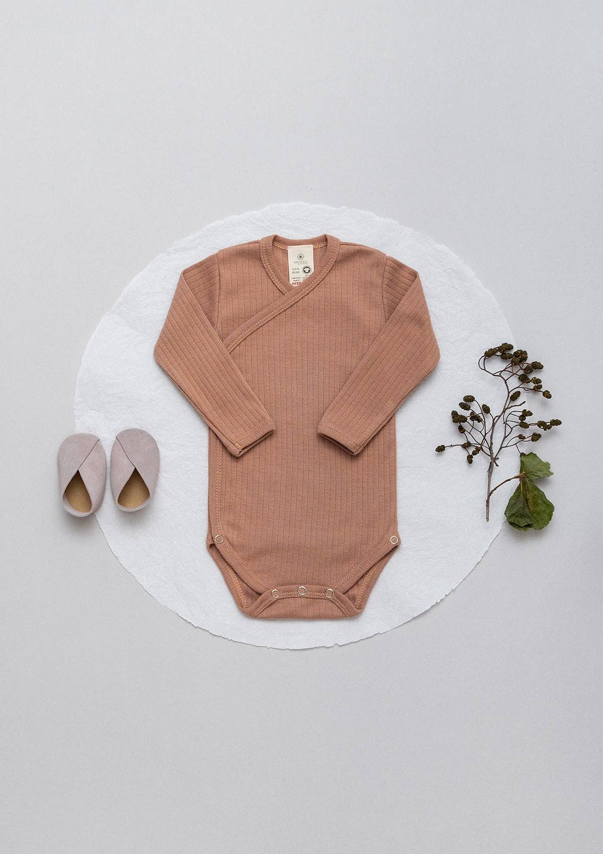 Wrap-style ribbed sienna onesie laid flat next to flowers and a pair of baby shoes