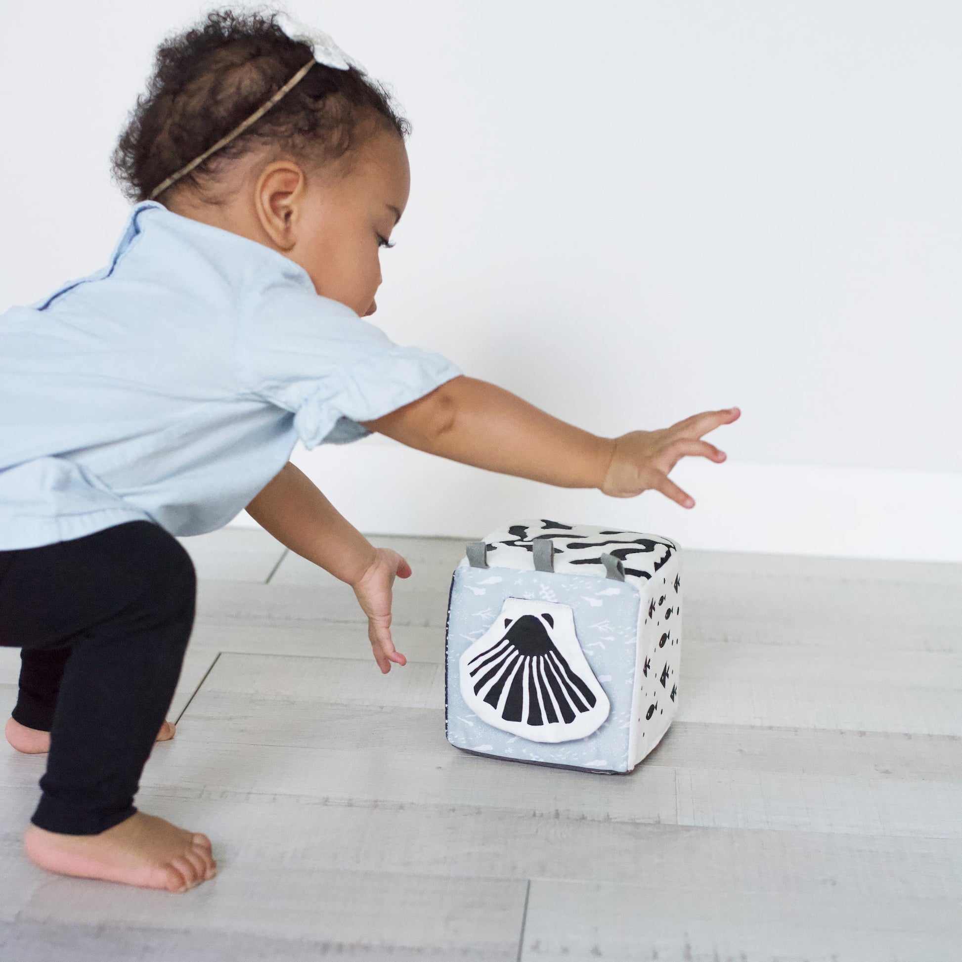 Child reaching towards a cube with a shell design on a light wooden floor.