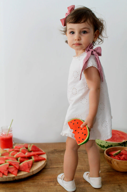 a child standing on a wooden table next to a plate of watermelon slices holding a natural rubber watermelon toy