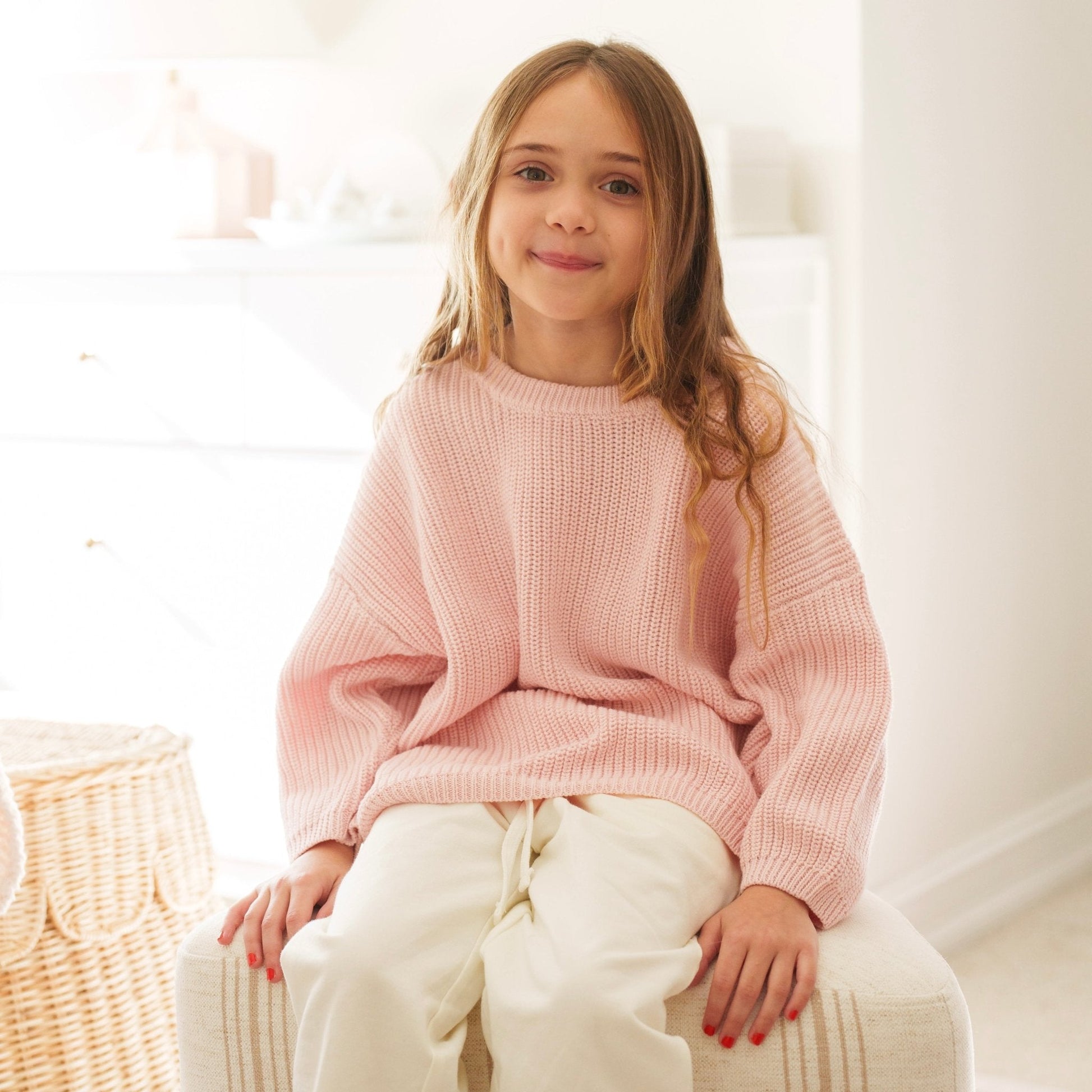 Young girl wearing a pink sweater sitting on a white chair.